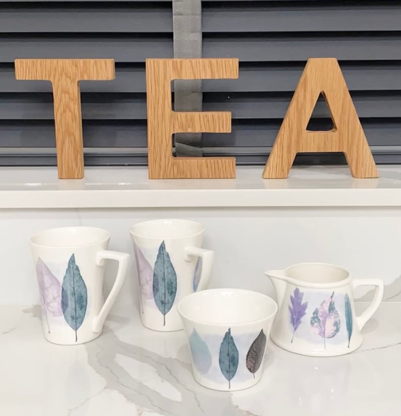 May include: A set of four ceramic mugs and a creamer with a leaf design in shades of purple, blue, and white. The mugs and creamer are on a white countertop with the word "TEA" spelled out in wooden letters.