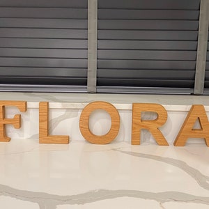 May include: Wooden letters spelling out the name "FLORA" in a light brown color. The letters are arranged in a row on a white countertop.