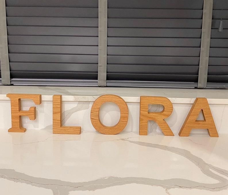 May include: Wooden letters spelling out the name "FLORA" in a light brown color. The letters are arranged in a row on a white countertop.