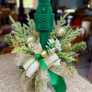 May include: A green velvet nutcracker figurine is the centerpiece of a holiday arrangement. The figurine is surrounded by faux evergreen branches, gold ornaments, and a decorative bow with green, white, and gold ribbons. The arrangement is festive and decorative.