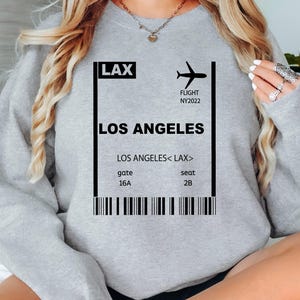 May include: A gray sweatshirt featuring a graphic of a boarding pass for Los Angeles (LAX). The pass includes the flight number NY2022, gate 16A, and seat 2B. The sweatshirt has a crew neck and long sleeves.