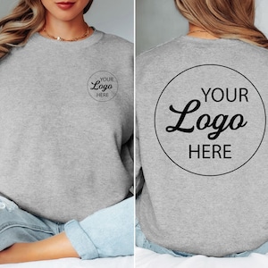 May include: A heather gray crewneck sweatshirt featuring a small circular black graphic on the left chest that reads YOUR LOGO HERE and a large circular black graphic on the back that reads YOUR LOGO HERE in a bold, centered font.