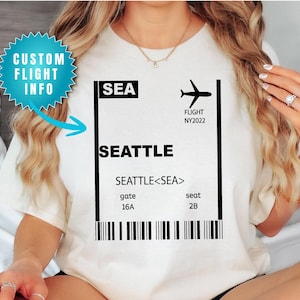 May include: Cream-colored t-shirt featuring a black and white graphic of a boarding pass for a flight to Seattle. The design includes the text "SEA", "SEATTLE", flight details, and a barcode. A turquoise starburst graphic reads "CUSTOM FLIGHT INFO".