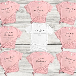 May include: Ten pink and white t-shirts with text that says "Bridesmaid", "Maid of Honor", "Team Bride", "Mother of the Bride", "The Bride", "Mother of the Groom", "Team Bride", "Chief Bridesmaid", and "Bridesmaid". Each shirt has a heart and a name printed on it.