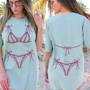 May include: A light blue t-shirt featuring a red bikini design on the front and back. The front design includes a bra and underwear with the text "Palm Springs 2026". The back design includes a bikini bottom with the word "Bridesmaid".