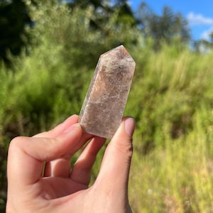 May include: A hand holding a polished, rectangular crystal point. The crystal is light brown and tan with a slightly translucent appearance. The background features green foliage and a blue sky.