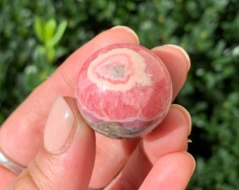 May include: A polished, spherical gemstone, primarily pink with concentric bands of white and a touch of gray-green at the base. The stone is held in a hand, showcasing its smooth, rounded form. The background is a blurred green, suggesting an outdoor setting.