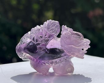 May include: A carved amethyst sculpture of a dragon-like creature with intricate details. The sculpture is a gradient of purple hues, with a detailed head, body, and legs. The dragon is posed on a white surface, casting a shadow.