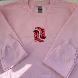 Puede incluir: Sudadera rosa con un par de botas de vaquero rojas bordadas en la parte delantera.