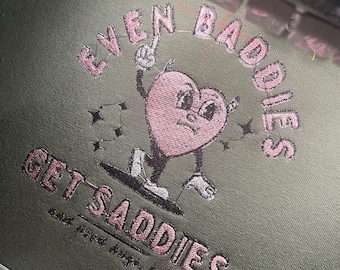 Puede incluir: Sudadera verde con texto bordado en rosa que dice "Even Baddies Get Saddies and food helps, lol". El bordado presenta un corazón rosa con cara y brazos, sosteniendo un signo de paz.