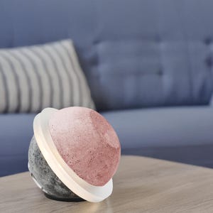 Può includere: Una moderna lampada sferica con un design bicolore. La metà superiore è di colore rosa polveroso, mentre la parte inferiore è grigio scuro. Un anello bianco illuminato circonda il centro. La lampada è appoggiata su una superficie di legno.