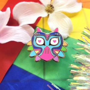 May include: A colorful enamel pin in the shape of an owl, featuring a gold outline and a vibrant design with blue, pink, and green accents. The owl has heart-shaped eyes and is set against a rainbow flag background.