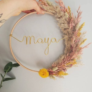 May include: A wooden hoop wreath decorated with dried flowers in shades of pink, yellow, and beige. The name "Maya" is spelled out in gold wire.