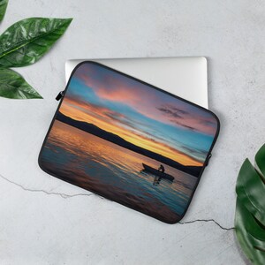 May include: A laptop sleeve with a colorful sunset over a lake with a silhouette of a person in a boat. The sky is a mix of pink, orange, blue, and purple.