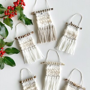 May include: Six handmade woven wall hangings with fringe, each attached to a small branch. The hangings are made with natural fibers in shades of white and beige.