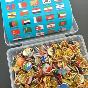 Könnte beinhalten: Eine transparente Box voller Reißzwecken mit Miniaturflaggen verschiedener Länder. Die Reißzwecken haben goldfarbene Köpfe. Im Hintergrund befindet sich eine blaue Box mit Länderflaggen.