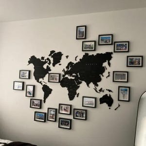 Peut inclure: Une carte du monde en bois noir avec un contour blanc des continents et des pays. La carte est montée sur un mur blanc et entourée de cadres photo noirs contenant des photos. Le mot "RUSSIA" est visible sur la carte.
