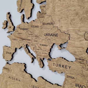 A wooden world map with the countries of Europe, Asia, and Africa labeled. The map is made of light brown wood with dark brown lines outlining the countries. The map is mounted on a white background.