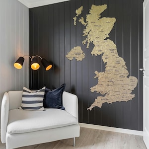 以下が含まれることがあります： イギリスとアイルランドの金色の木製地図が、濃い灰色の壁に取り付けられています。地図には郡名が詳細に記載されています。地図の下には、ストライプと無地の青い枕が付いた白いアームチェアがあります。左側には、3つのライトが付いた黒いランプがあります。