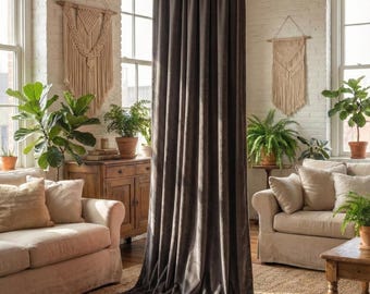 May include: A dark brown velvet curtain in a living room with beige sofas, plants, and macrame wall hangings. The curtain is long and drapes to the floor, creating a cosy atmosphere. The room has natural light from large windows.