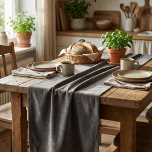 Könnte beinhalten: Ein rustikaler Holztisch, der für eine Mahlzeit gedeckt ist. Ein grauer Tischläufer erstreckt sich über die Länge des Tisches, mit Tellern, Tassen und Servietten für jeden Platz. Ein Brotkorb und Topfpflanzen tragen zur gemütlichen Atmosphäre bei.