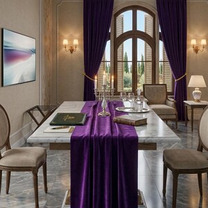 Puede incluir: Un comedor con una mesa de mármol adornada con un camino de mesa de terciopelo morado oscuro. Un candelabro plateado y cristalería están dispuestos. La habitación cuenta con ventanas arqueadas con contraventanas, cortinas de terciopelo morado y apliques de pared.