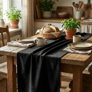 Puede incluir: Una mesa de comedor rústica de madera preparada para una comida. Un camino de mesa negro se extiende por el centro, con platos, tazas y una cesta de pan. Las plantas en macetas y una ventana contribuyen al ambiente acogedor.