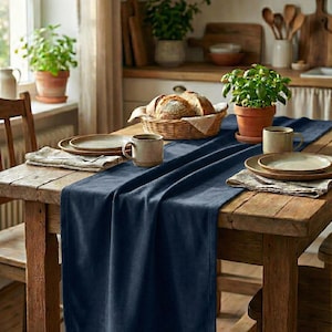 Può includere: Un tavolo da pranzo rustico in legno è apparecchiato con un runner blu navy, piatti, tazze e un cestino di pane. Erbe in vaso e una finestra con luce naturale completano la scena. Il tavolo è in legno.