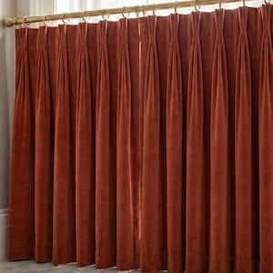 Op de afbeelding: Roestkleurige fluwelen gordijnen die aan een goudkleurige stang met ringen hangen. De gordijnen zijn geplooid en hangen in verticale plooien, wat een rijke, getextureerde uitstraling creëert. De gordijnen bevinden zich voor een raam.