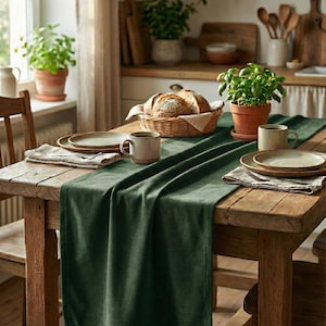 Puede incluir: Una mesa de comedor rústica de madera está decorada con un camino de mesa verde oscuro, platos, tazas y servilletas de lino. Una cesta de pan y plantas en macetas contribuyen a la atmósfera acogedora y acogedora. La escena está bañada por la luz natural.