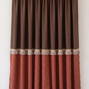 Peut inclure: Un rideau pleine longueur composé d'une section supérieure marron foncé et d'une section inférieure terracotta, séparées par une frange beige. Le rideau présente une tête à plis pincés et est suspendu à une tringle argentée.
