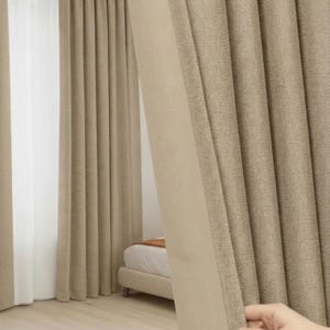 Op de afbeelding: Beige gordijnen met textuur en een lichtgekleurde rand worden gedeeltelijk teruggetrokken, waardoor een glimp van een slaapkamer zichtbaar wordt. De gordijnen hangen voor een wit, transparant gordijn. De kamer heeft een lichtgekleurde houten vloer.