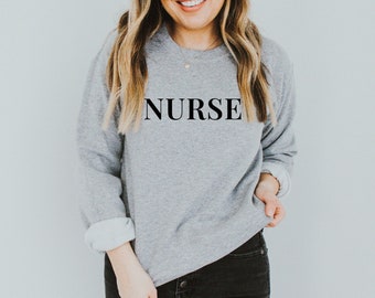 Puede incluir: Una sudadera de cuello redondo y manga larga en color gris jaspeado con la palabra NURSE impresa en letras mayúsculas negras en el pecho. La prenda tiene cuello y puños acanalados, con las mangas remangadas hasta los codos, combinada con pantalones vaqueros negros.