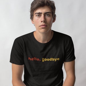 Puede incluir: Una camiseta negra de manga corta con el texto "hello, goodbye" impreso en el pecho. La palabra "hello" es de un tono rojo anaranjado cálido, mientras que la palabra "goodbye" es de un color amarillo dorado contrastante, ambas en fuente simple.