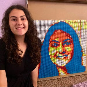 以下が含まれることがあります： カラフルな正方形のタイルで作られた大きなモザイク肖像画の前に人が立っています。肖像画は、長い黒髪と明るい笑顔の人物です。