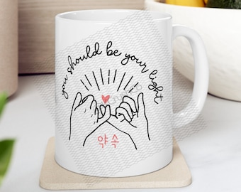 Könnte beinhalten: Weiße Keramiktasse mit einer Schwarzweißillustration von zwei Händen, die sich einen kleinen Finger versprechen. Der Text "you should be your own light" ist in einem Kreis um die Hände geschrieben. Das koreanische Wort "약속" steht unter den Händen.