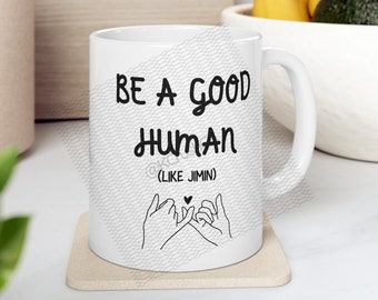 Könnte beinhalten: Weiße Keramiktasse mit schwarzem Text, der "Sei ein guter Mensch (wie Jimin)" lautet, und einer schwarzen Illustration von zwei Händen, die ein Pinky-Versprechen machen.