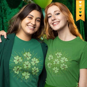 以下が含まれることがあります： 緑色のTシャツを着た2人の女性。各クローバーには名前が書かれています。シャツは無地の緑色です。背景には緑色の風船と緑色のカーテンがあります。