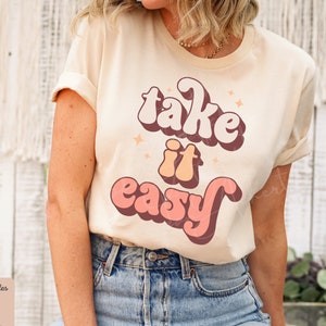 Könnte beinhalten: Ein weißes T-Shirt mit dem Text "take it easy" in einer Retro-Schriftart in bunten Farben. Der Text ist in den Farben Rosa, Orange und Braun gehalten.