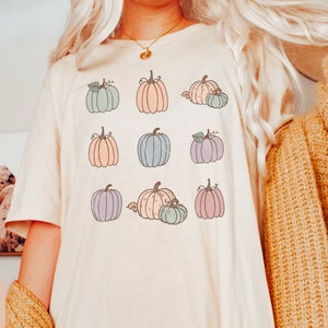 May include: A white t-shirt with a pattern of pastel colored pumpkins. The pumpkins are arranged in a grid pattern and have different shapes and sizes.