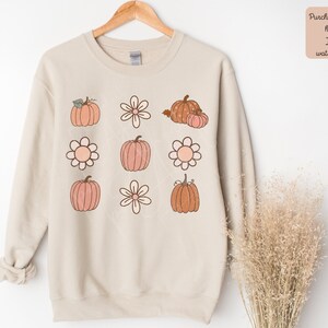 Op de afbeelding: Een beige sweatshirt met een grafisch ontwerp van meerdere oranje en bruine pompoenen en witte bloemen. Het ontwerp wordt in een raster herhaald.