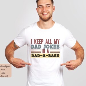 Könnte beinhalten: Weißes T-Shirt mit einer Grafik im Retro-Stil, auf dem "I keep all my dad jokes in a dad-a-base" steht.