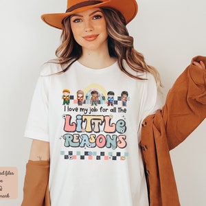 Pu&ograve; includere: Maglietta bianca con una grafica colorata che dice "I love my job for all the little reasons". La grafica presenta un arcobaleno, uno schema a scacchi e bambini dei cartoni animati.
