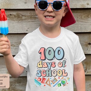 K&ouml;nnte beinhalten: Ein wei&szlig;es T-Shirt mit dem Text "100 Tage Schule" in bunten, Retro-Lettern. Das Shirt zeigt verschiedene schulthemenbezogene Illustrationen, darunter einen Bleistift, einen Pinsel, einen Globus und ein Lineal.