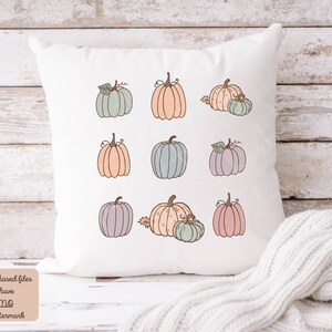 May include: A white pillow with nine pastel colored pumpkins with brown stems and green leaves. The pumpkins are arranged in three rows of three. The pillow is sitting on a light wood surface.