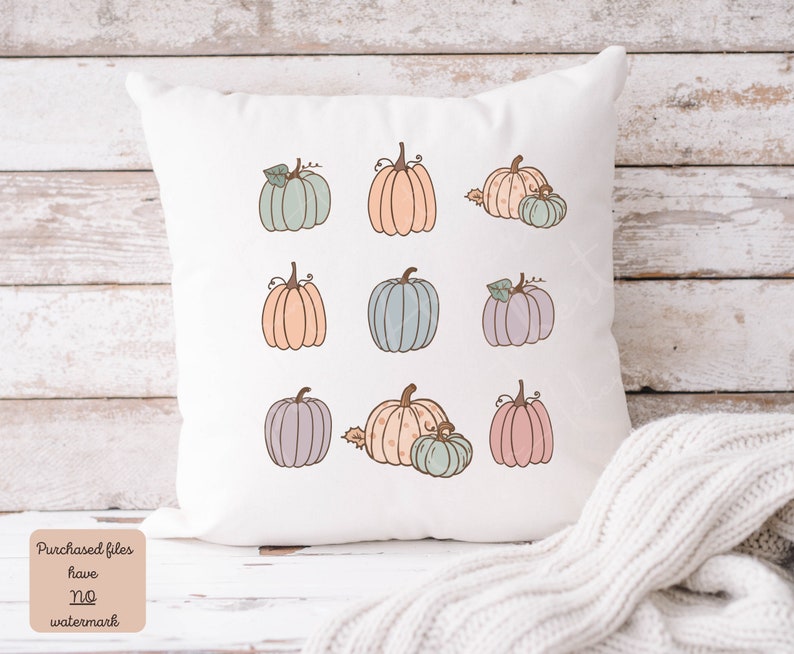 May include: A white pillow with nine pastel colored pumpkins with brown stems and green leaves. The pumpkins are arranged in three rows of three. The pillow is sitting on a light wood surface.