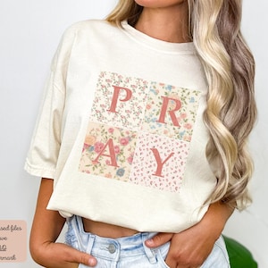 May include: A white t-shirt with a floral print design that spells out the word "PRAY" in pink letters.