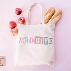 May include: A white canvas tote bag with the word "MIDWIFE" printed in colorful, block letters. The bag is filled with two loaves of bread and red apples. The bag is on a pink background.