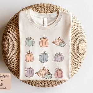 May include: A beige t-shirt with a design of nine pastel-colored pumpkins. The pumpkins are arranged in three rows of three. The pumpkins are all different shapes and sizes. The t-shirt is on a woven straw surface.