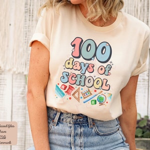K&ouml;nnte beinhalten: Ein beiges T-Shirt mit einer bunten Grafik, auf der "100 Tage Schule" mit Schulutensilien und Sternen steht.